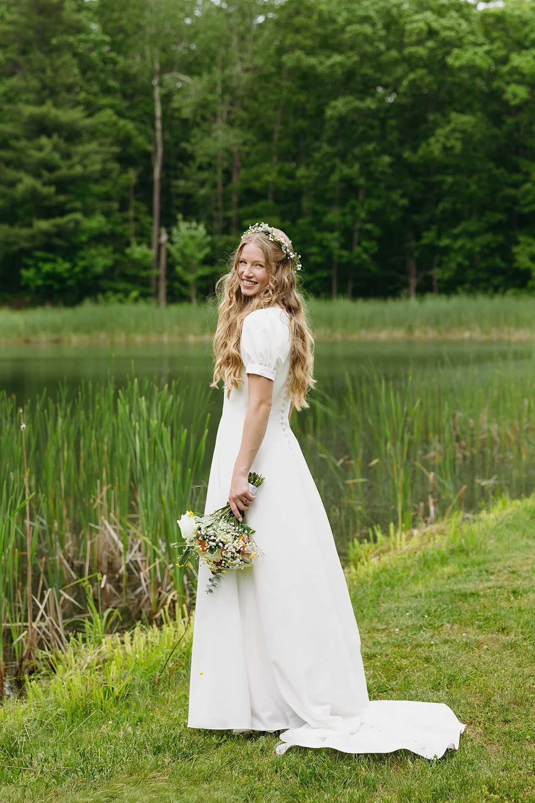 Woman in a white dress with puff sleeves and a flower crown smiles while standing outdoors on green grass for a backyard wedding