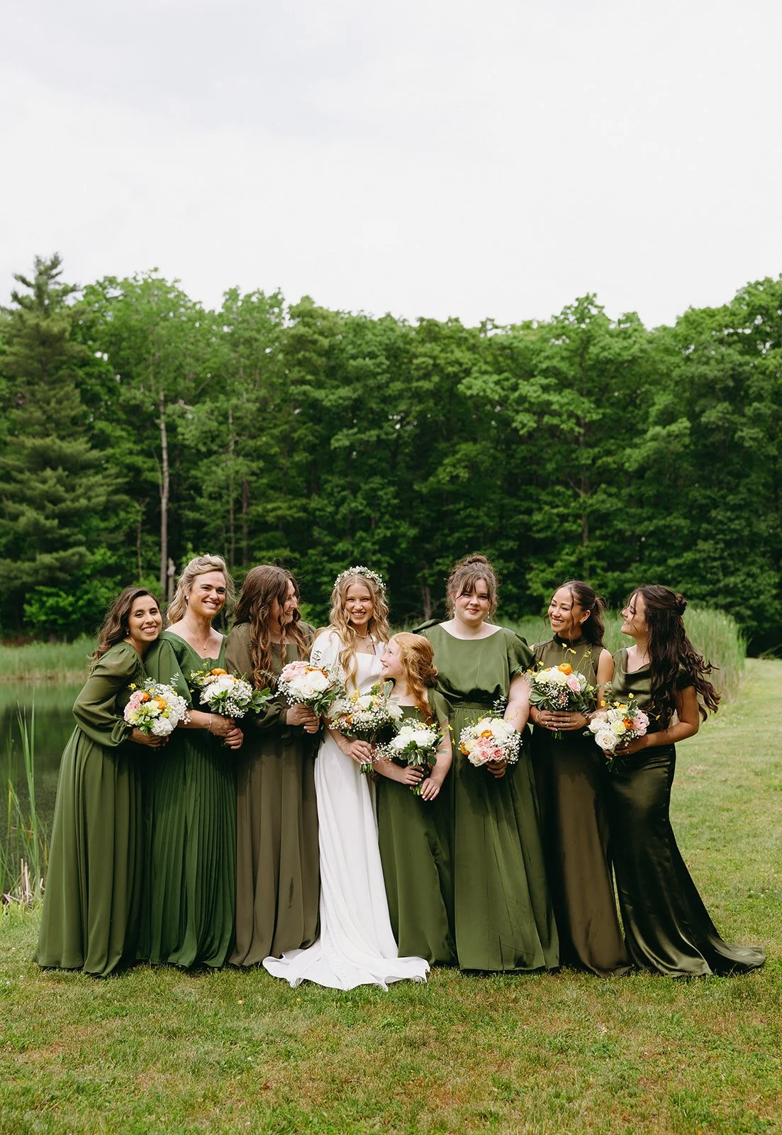 Bride in white dress stands with six bridesmaids in various shades of green dresses, all holding bouquets, outdoors near trees and a pond.