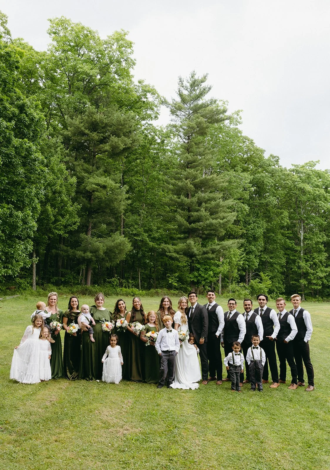 A wedding party poses outdoors in formal attire, with bridesmaids in green dresses and groomsmen in black vests, surrounded by green trees.