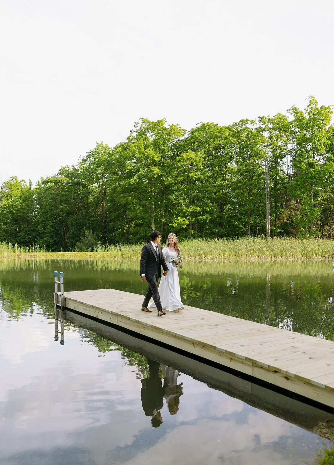 A couple in formal attire walks together on a wooden dock over a calm lake, with green trees and reeds in the background.