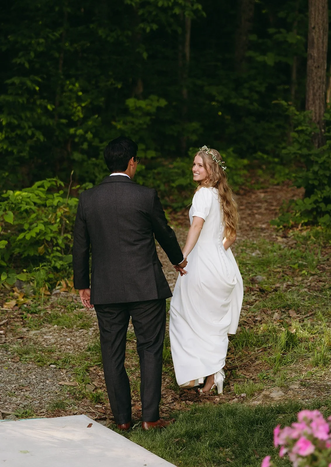 A bride in a white dress and groom in a dark suit walk hand in hand along a wooded path, facing away from the camera.