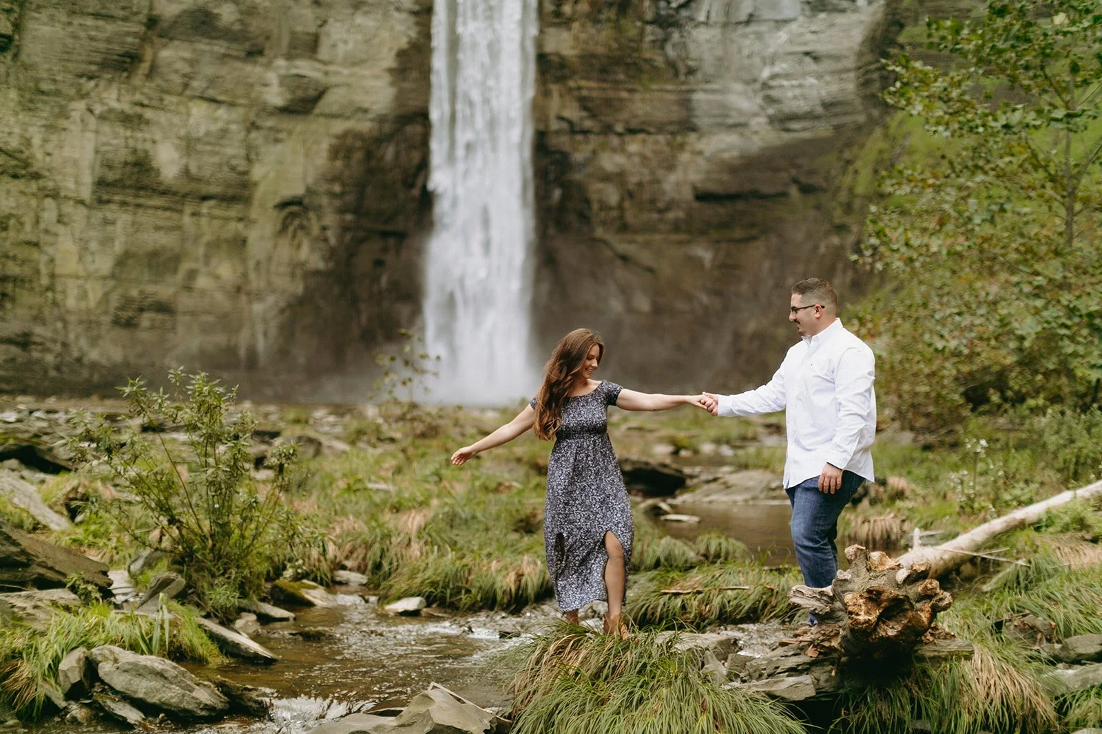 A couple walks toward a tall waterfall cascading down a sheer rock cliff.