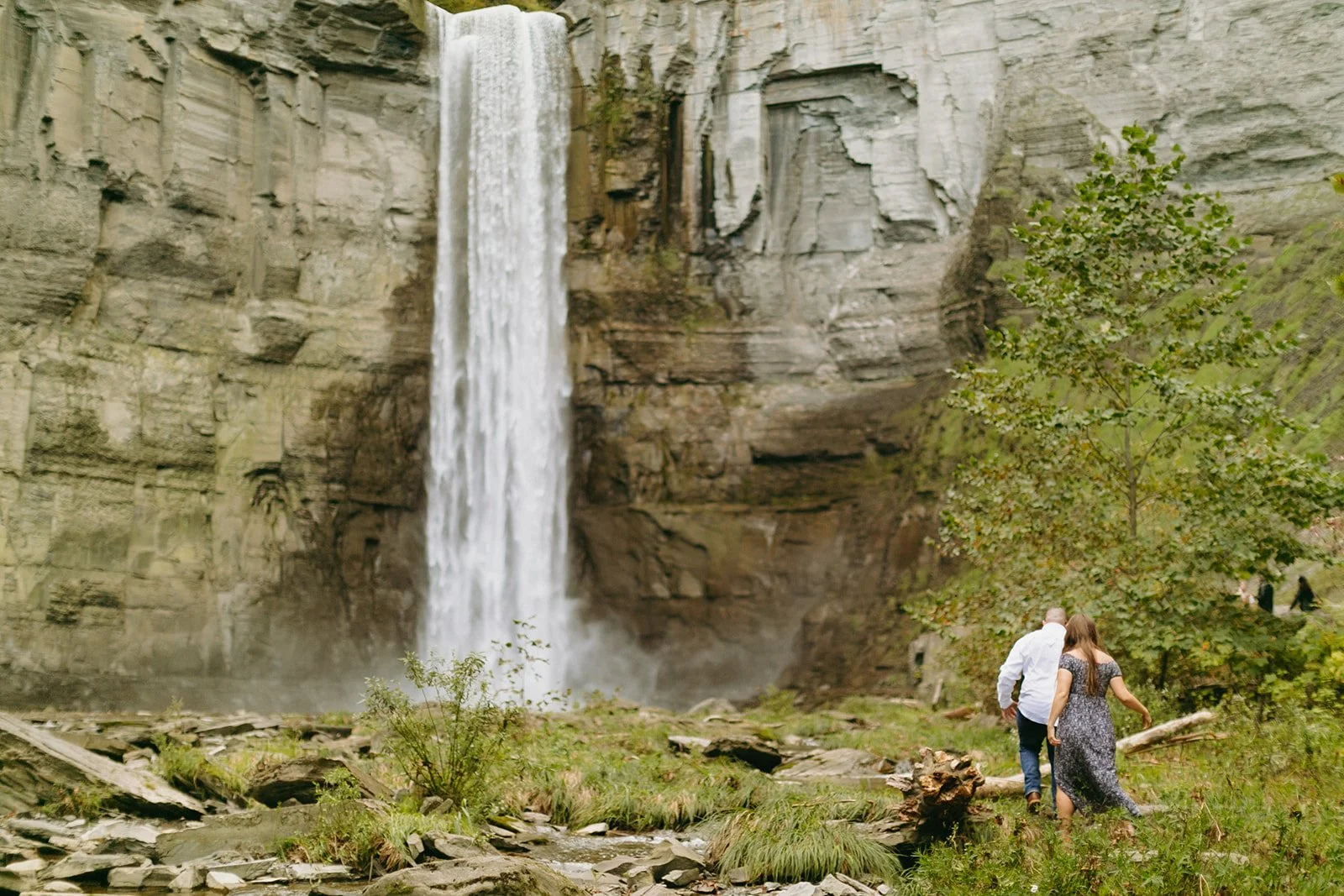 A couple walks toward a tall waterfall cascading down a sheer rock cliff.