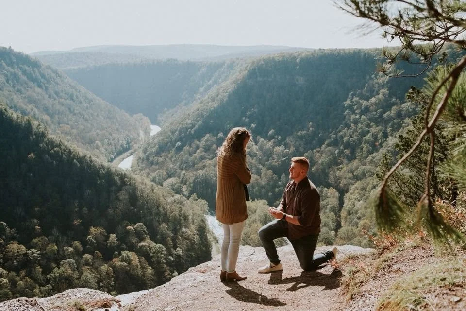 A couple standing on the edge of a cliff overlooking a forested valley.
