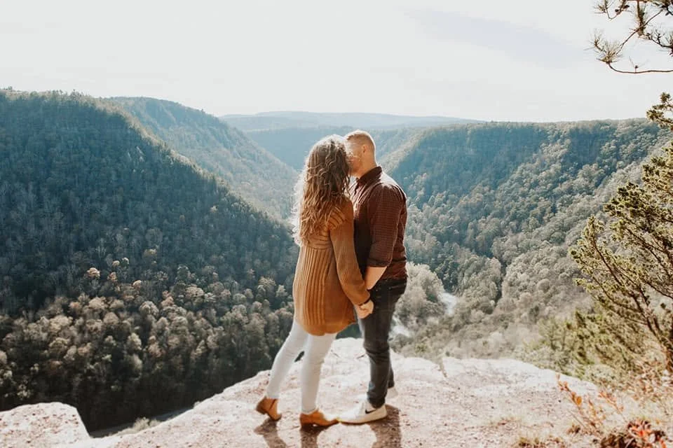 A couple standing on the edge of a cliff overlooking a forested valley.