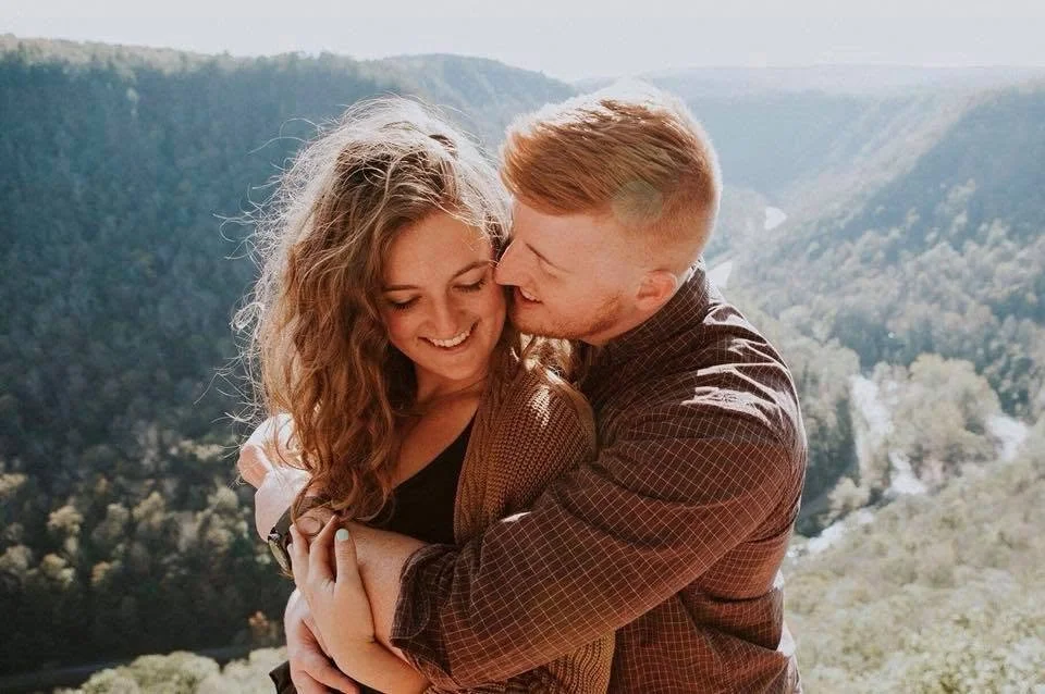 A couple standing on the edge of a cliff overlooking a forested valley.