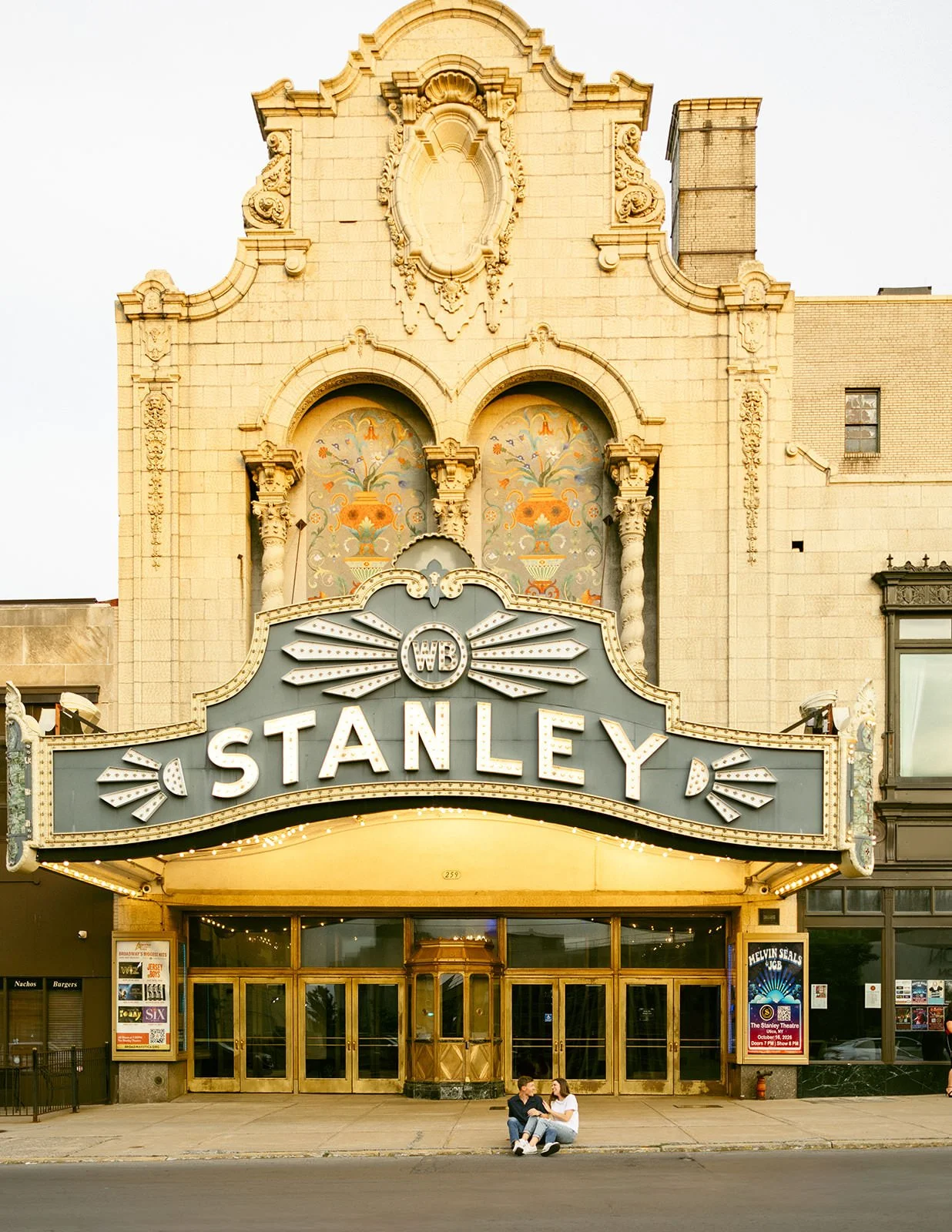 Engaged couple sitting together beneath the marquee of the historic Stanley Theatre in downtown Utica, NY, surrounded by the theater’s ornate architecture and vintage signage.
