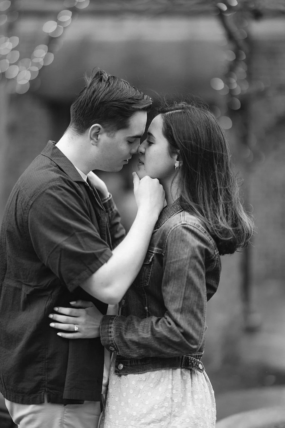 Black and white photo of a man gently holds a woman’s chin as he leans in for a kiss during their engagement photo, capturing an intimate and romantic moment outdoors.
