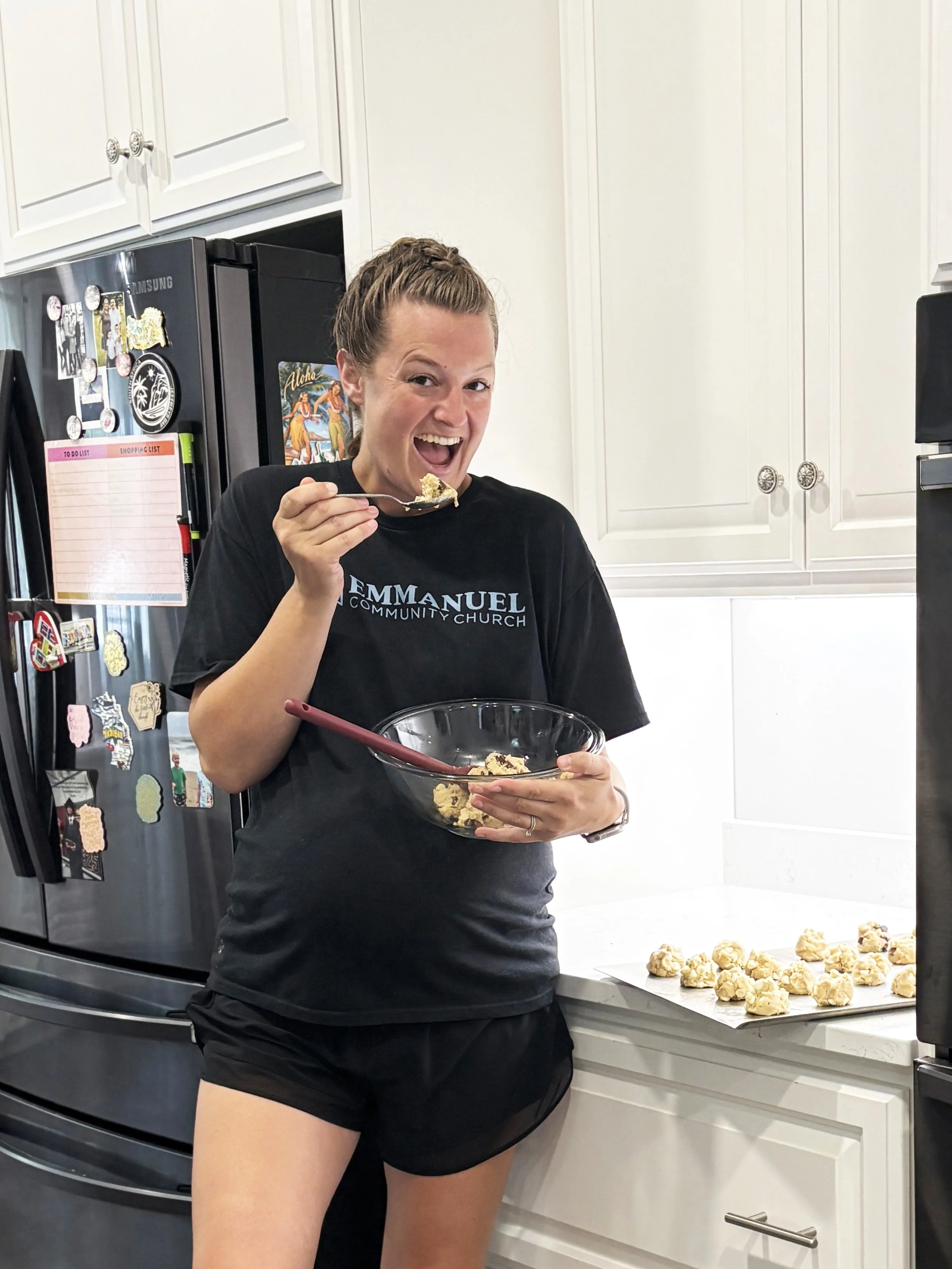 Pregnant woman in black t-shirt baking and eating cookie dough in bright white kitchen.