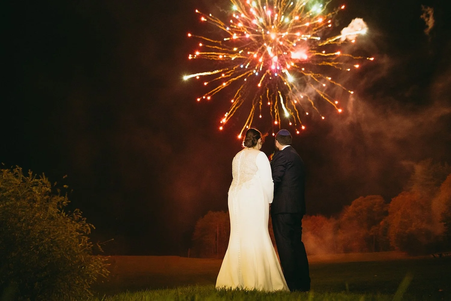 The bride and groom stand close together observing the fireworks the light up the sky.