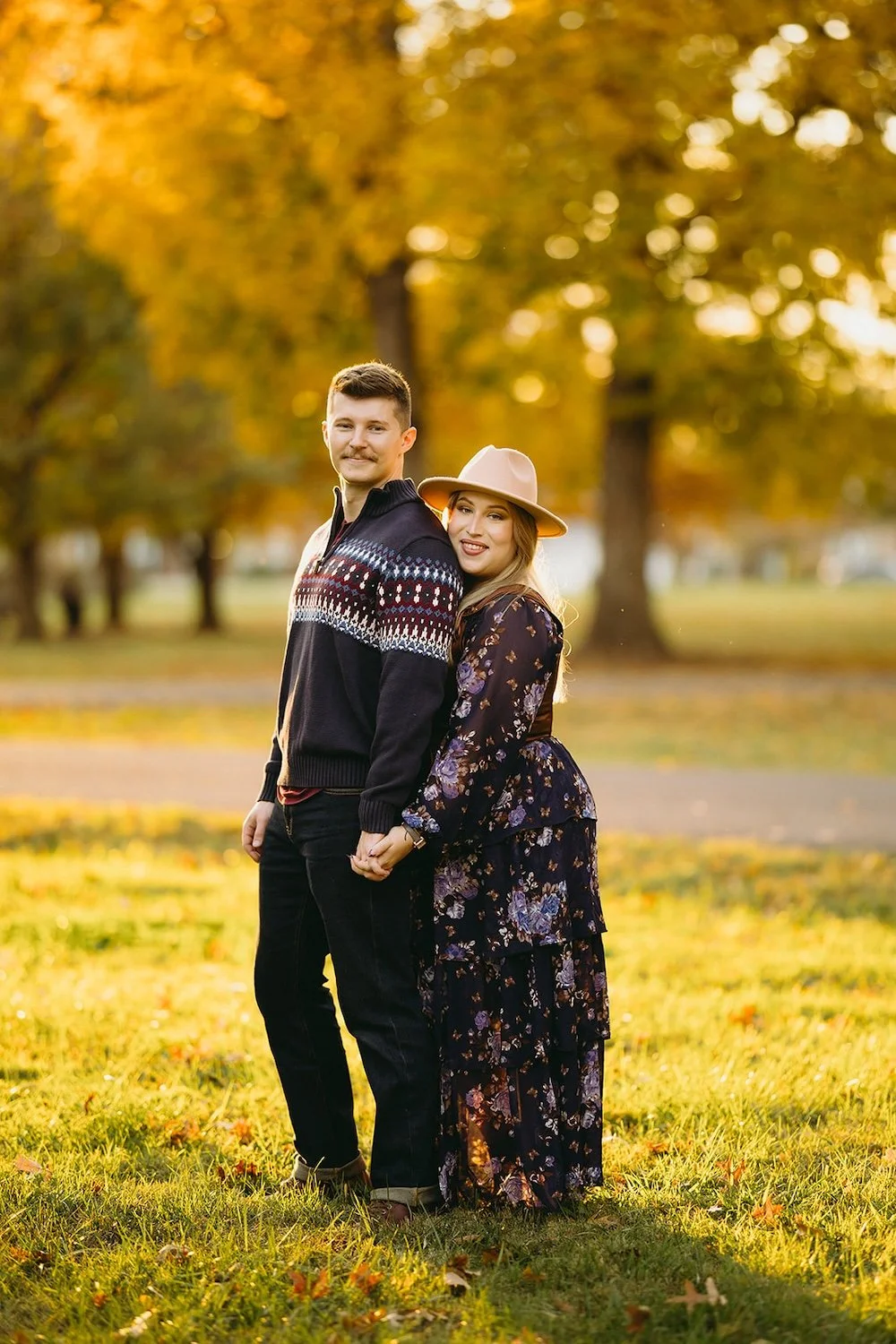 The couple stand posing in the park surrounded by bright sun and fall colors.