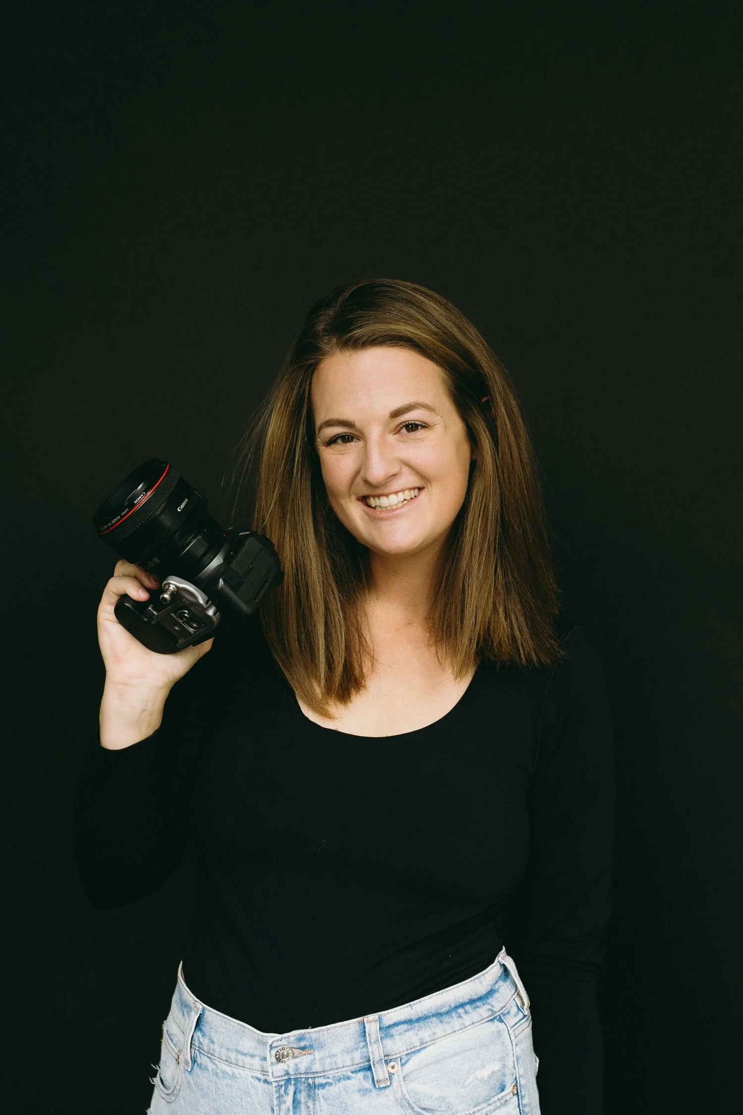 Professional headshot of a smiling wedding photographer holding a camera against a dark backdrop, authentic and friendly brand imagery.