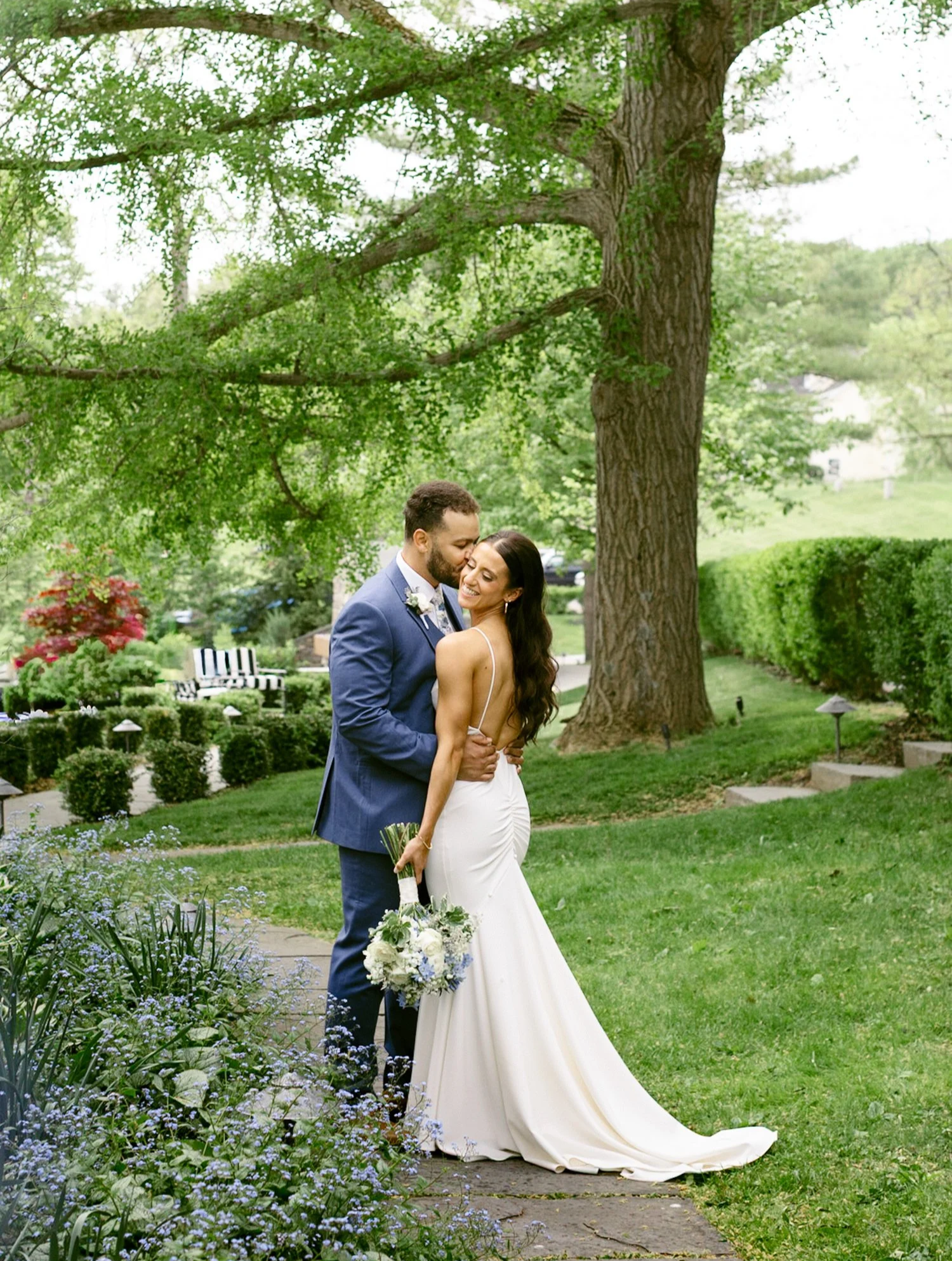 Bride and groom embracing in a lush garden beneath a large tree, romantic outdoor wedding photography.