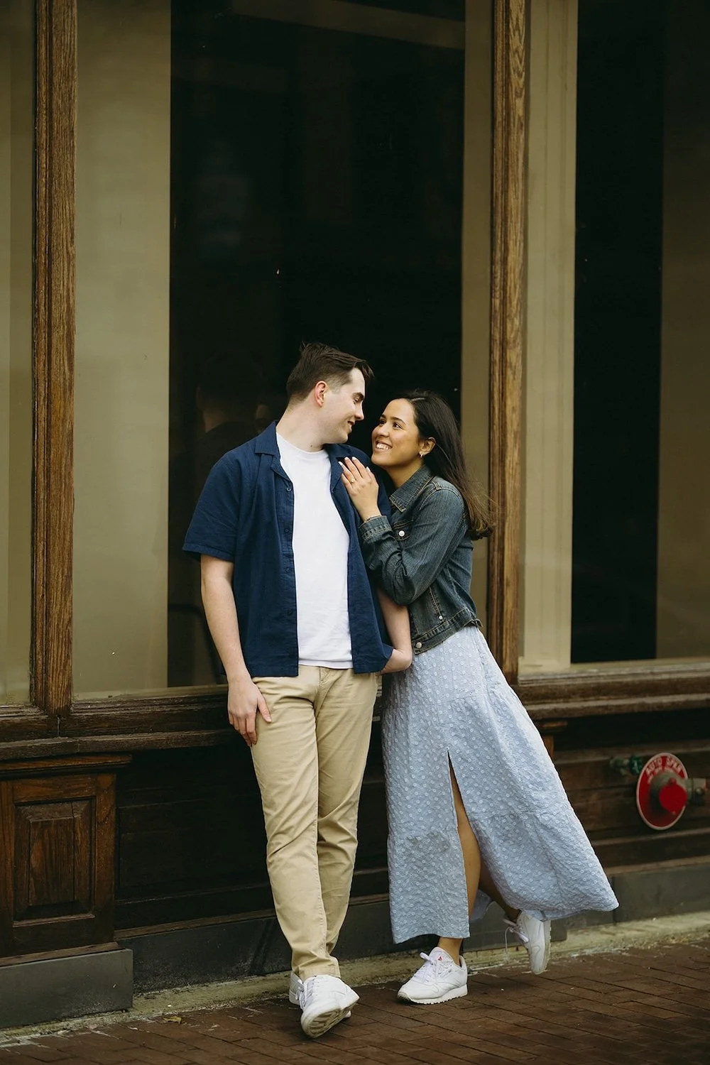The couple lean against building as the future bride looks into her grooms eyes.