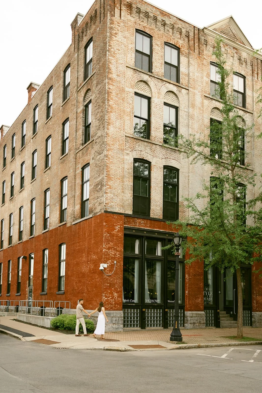 Engaged couple holding hands while walking past a historic brick building in downtown Utica, NY, surrounded by tall black-framed windows, a leafy tree, and vintage architecture.