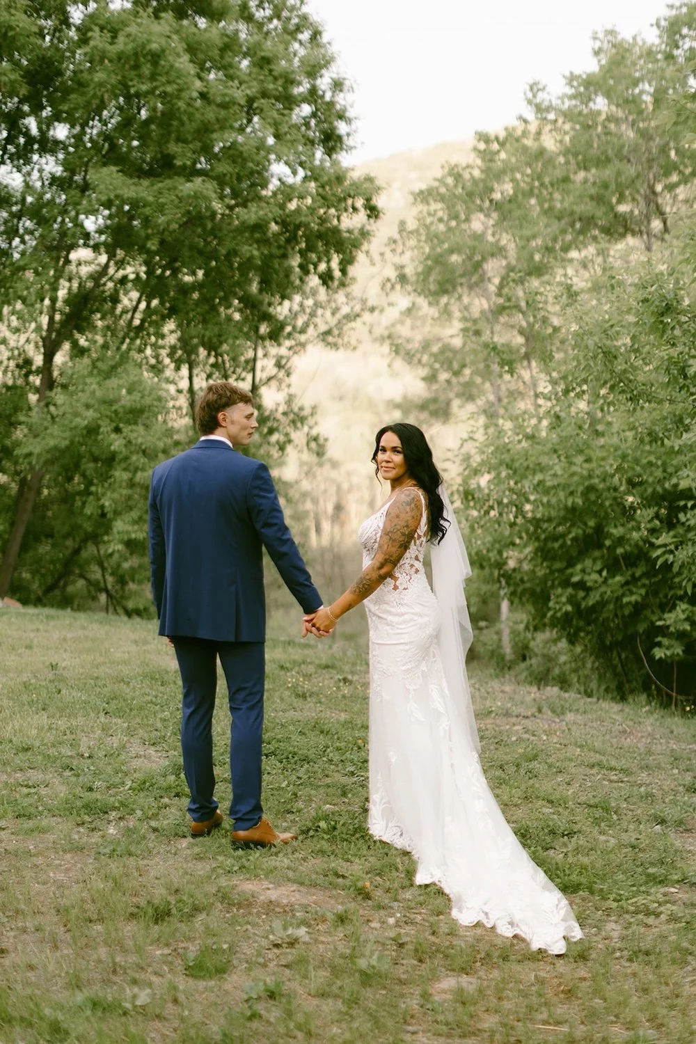 Bride and groom walk together through the lush greenery hand in hand. The bride looks back.