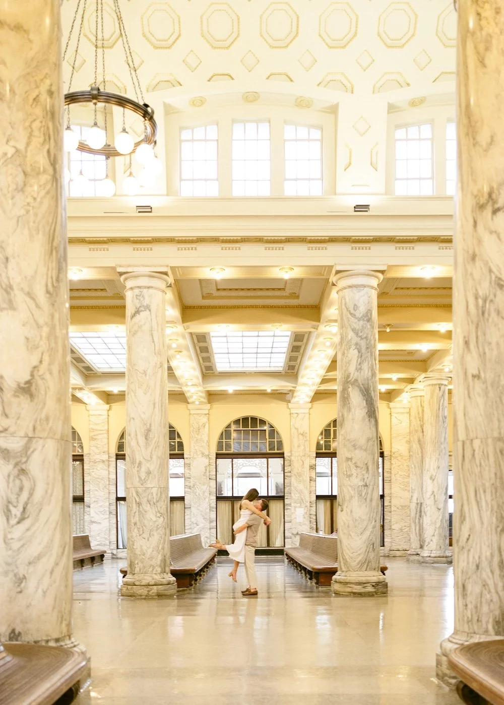 Couple embracing inside historic Union Station in Utica, New York, surrounded by marble columns and grand architecture.