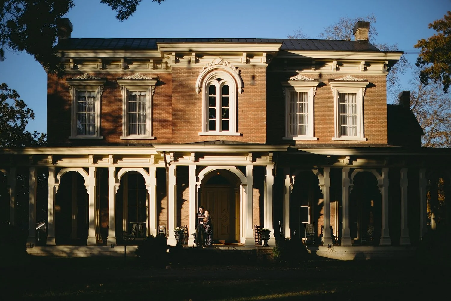The couple stand on the porch of historic home leaning against post holding one another.