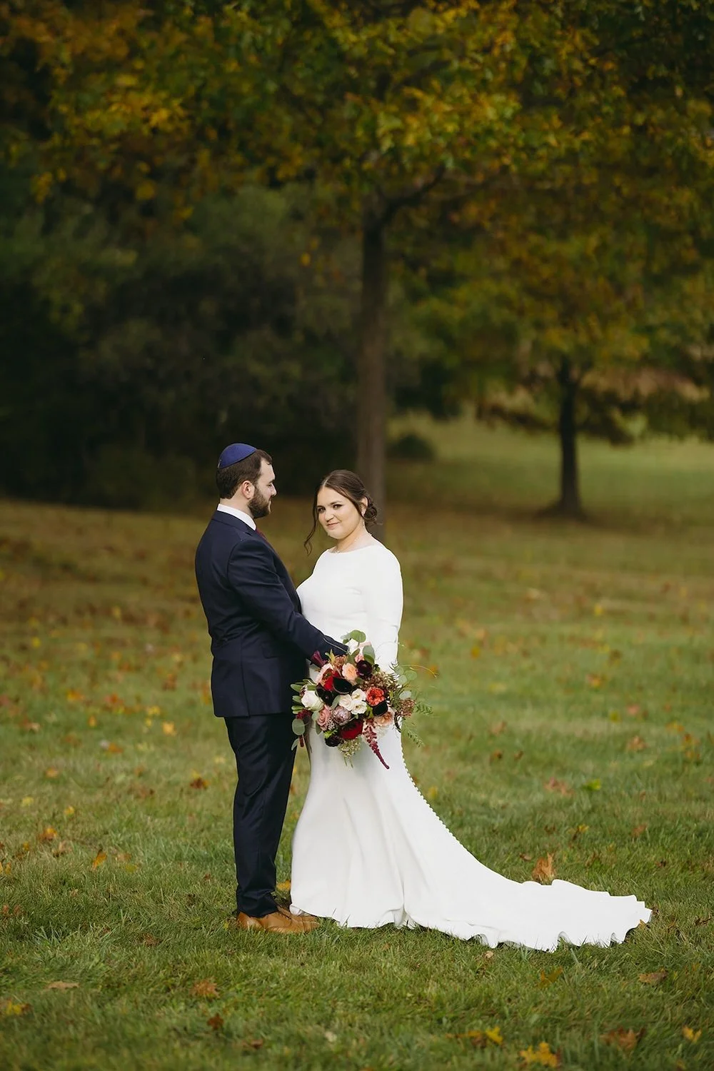 The bride and groom stand in the field decorated with trees as the groom admires his bride. Bride looks toward the camera.