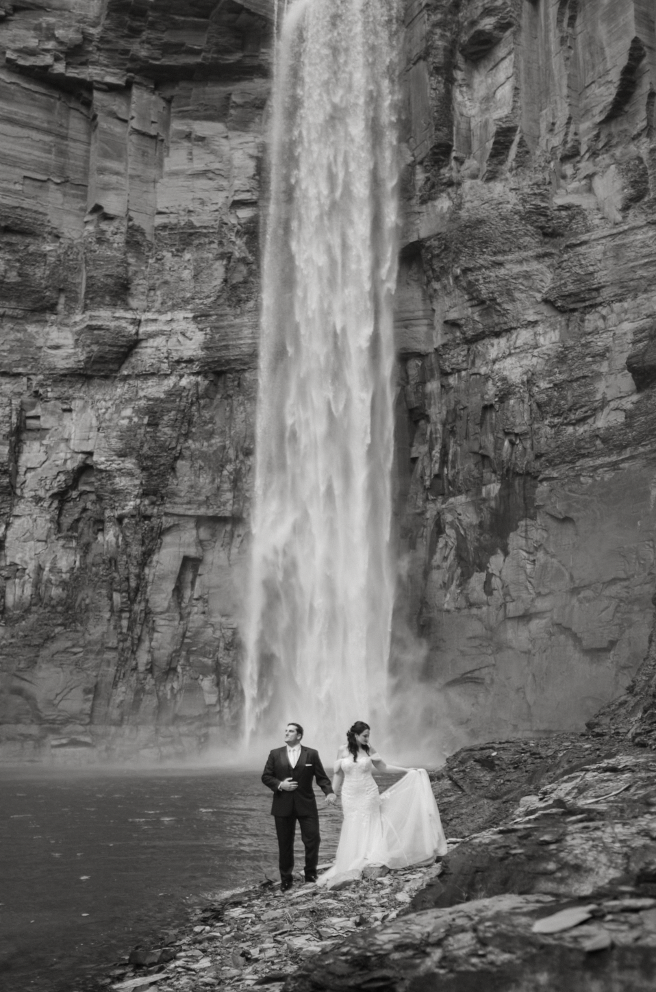 Black and white wedding portrait of a bride and groom standing beneath a dramatic waterfall, adventurous upstate new york wedding photography.