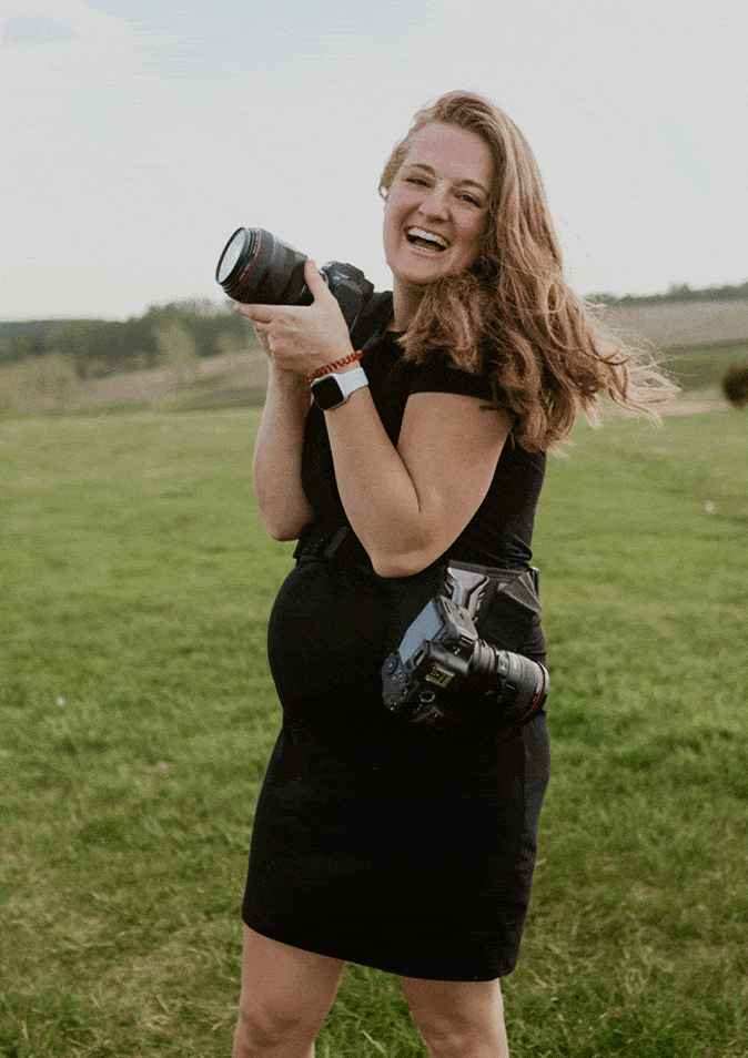 Laughing female wedding photographer holding cameras in grassy outdoor wedding venue.