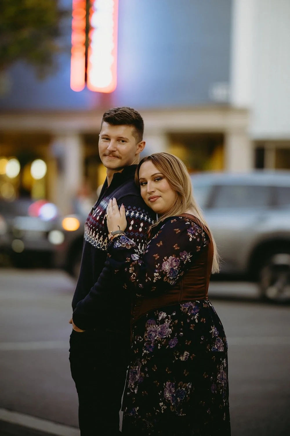 The couple stand close together in downtown looking both at the camera as the future bride holds her groom close.