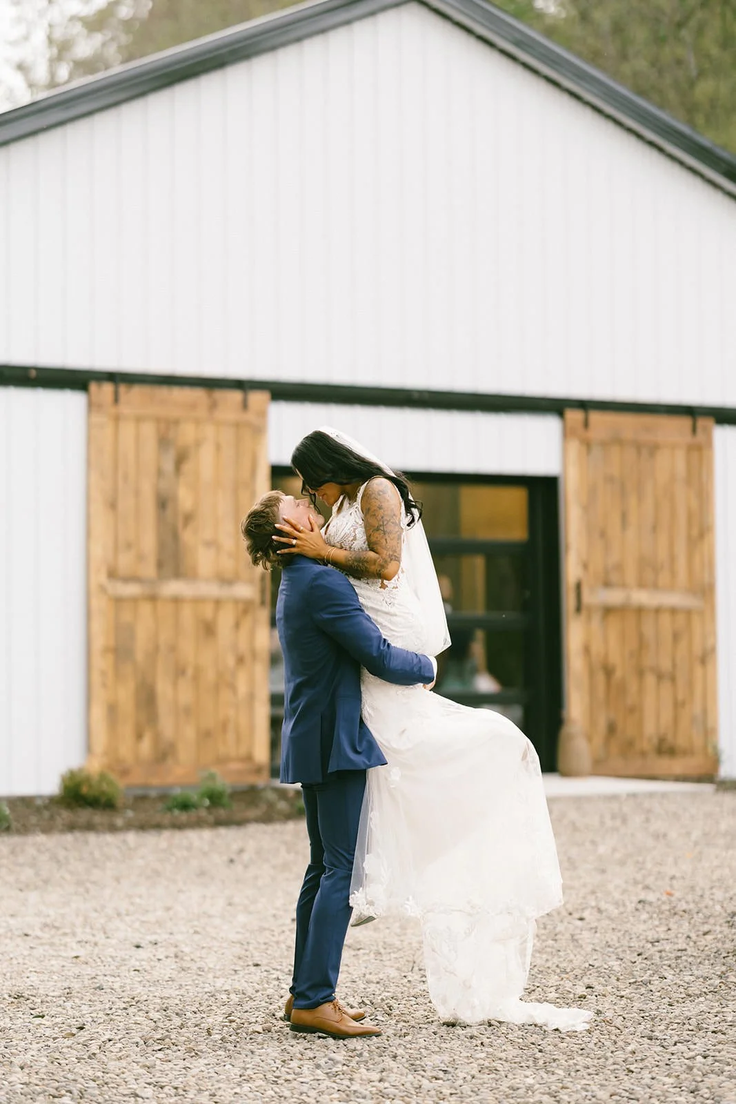Bride and groom sharing a joyful lift in front of a white barn venue, rustic outdoor wedding photography in Upstate New York.