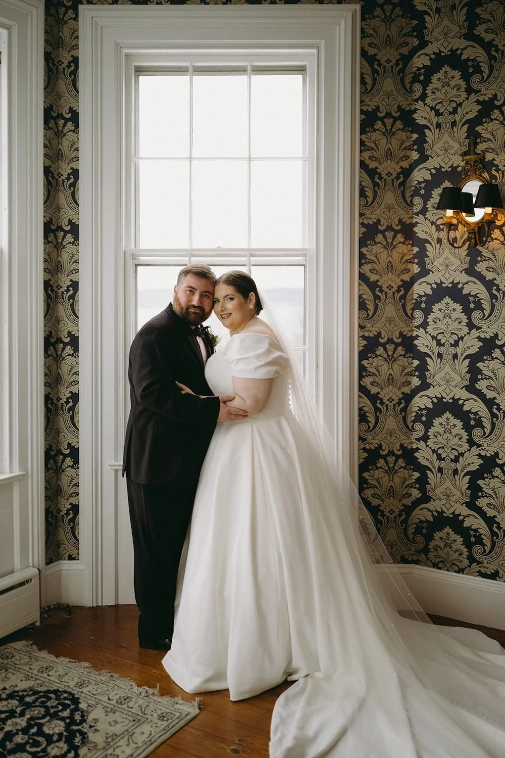 The couple stand holding one another as their heads touch posing in front of a window.