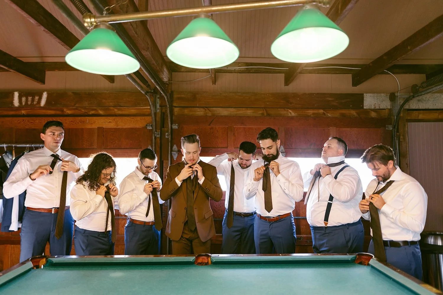 The groomsmen stand in front of the pool table while they tie their ties.