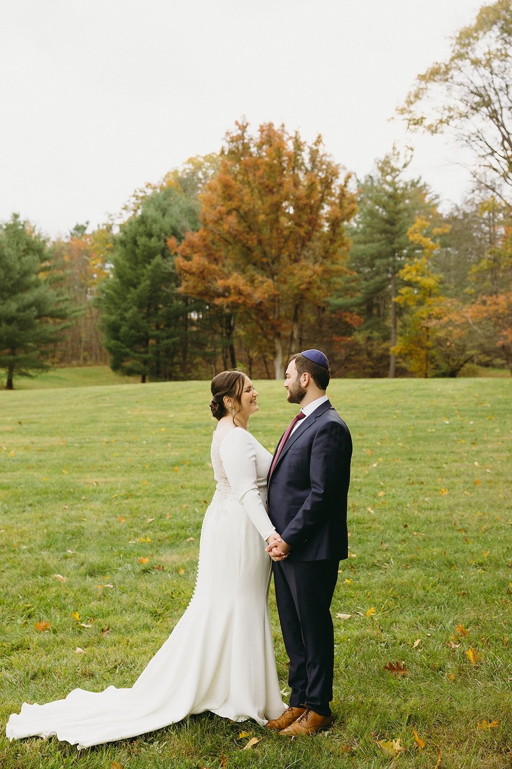 The couple stand facing each other smiling with the fall foliage decorated behind them.