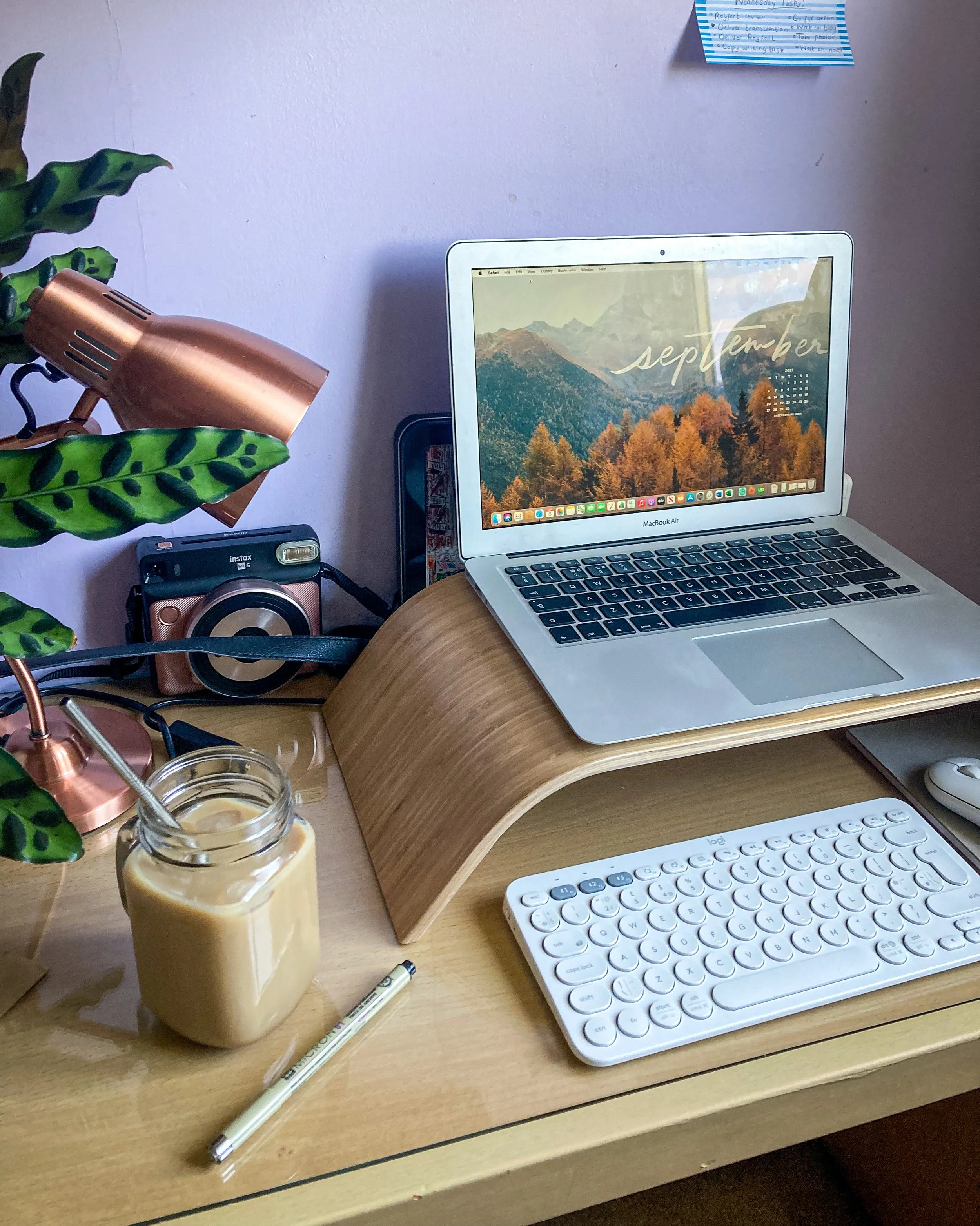 A desk shot featuring a laptop computer on a stand with a white keyboard underneath. There's a cup of iced coffee in a mason jar, a Polaroid camera, a pen, a plant and a desk lamp.
