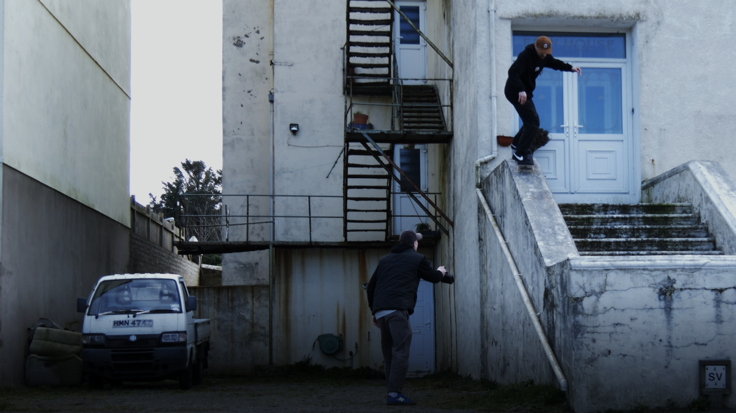 Two people are skateboarding outside near an old, worn building with stairs and a blue door.