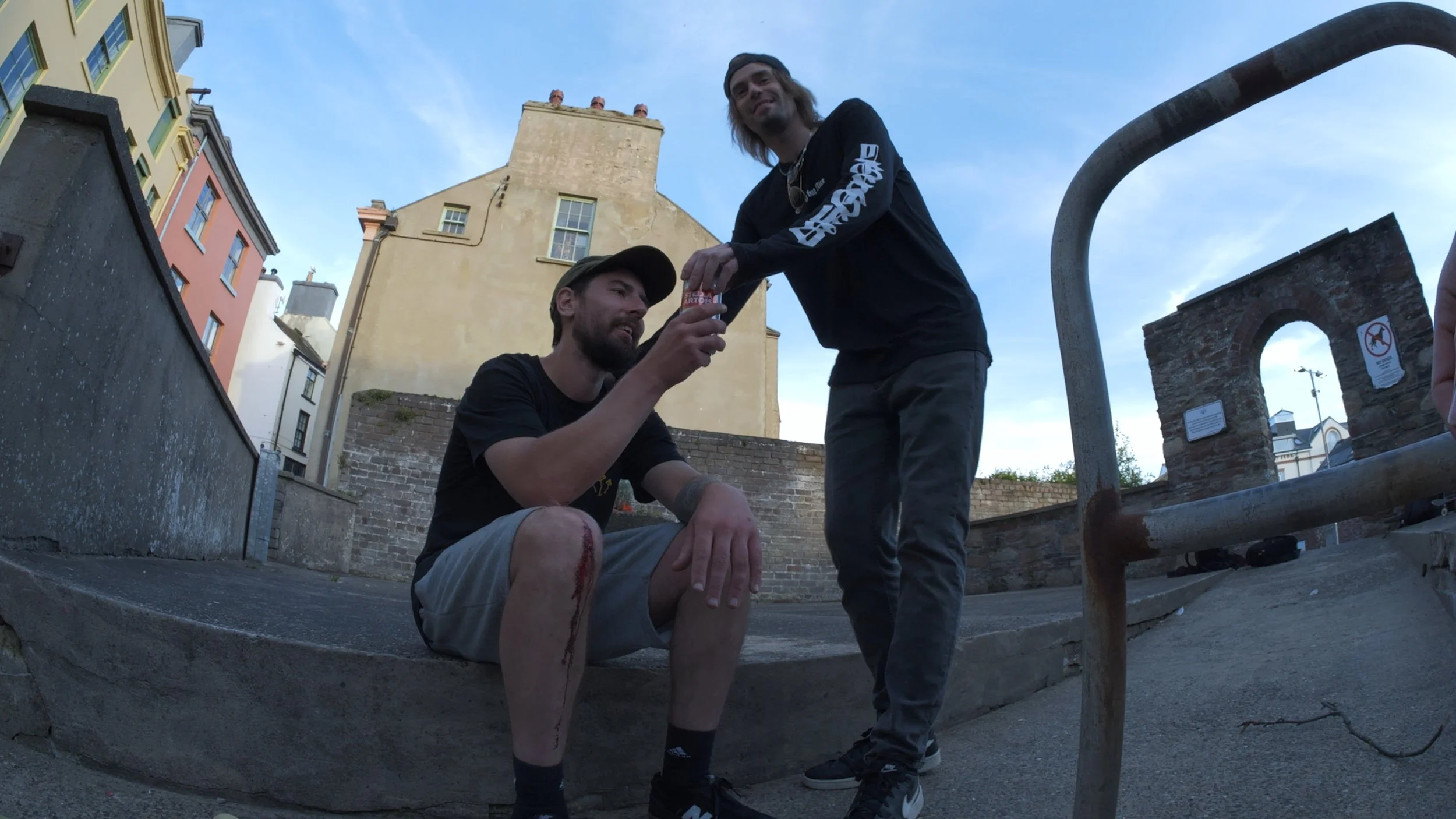 Two men outdoors, one sitting on the sidewalk with a leg injury and blood visible, the other standing and handing him a drink, with buildings and a blue sky in the background.