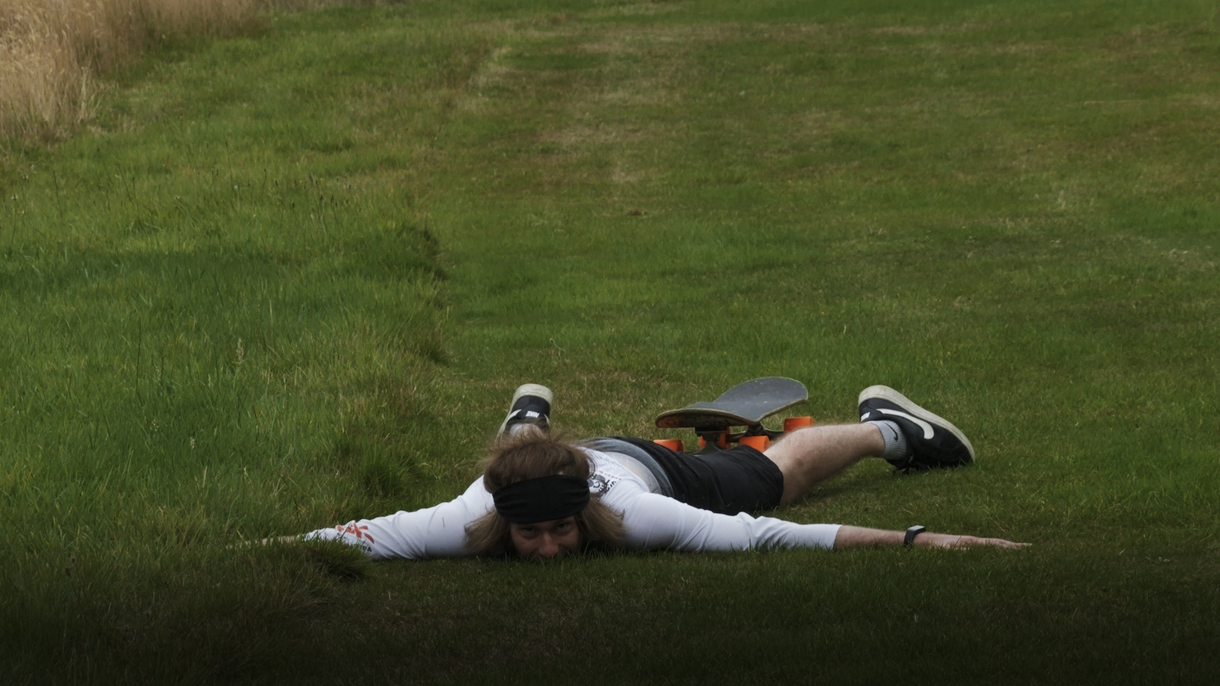 Person lying face down on grass with arms and legs extended, wearing a white long-sleeve shirt, black shorts, black sneakers, and a black headband, with a skateboard resting on their back.