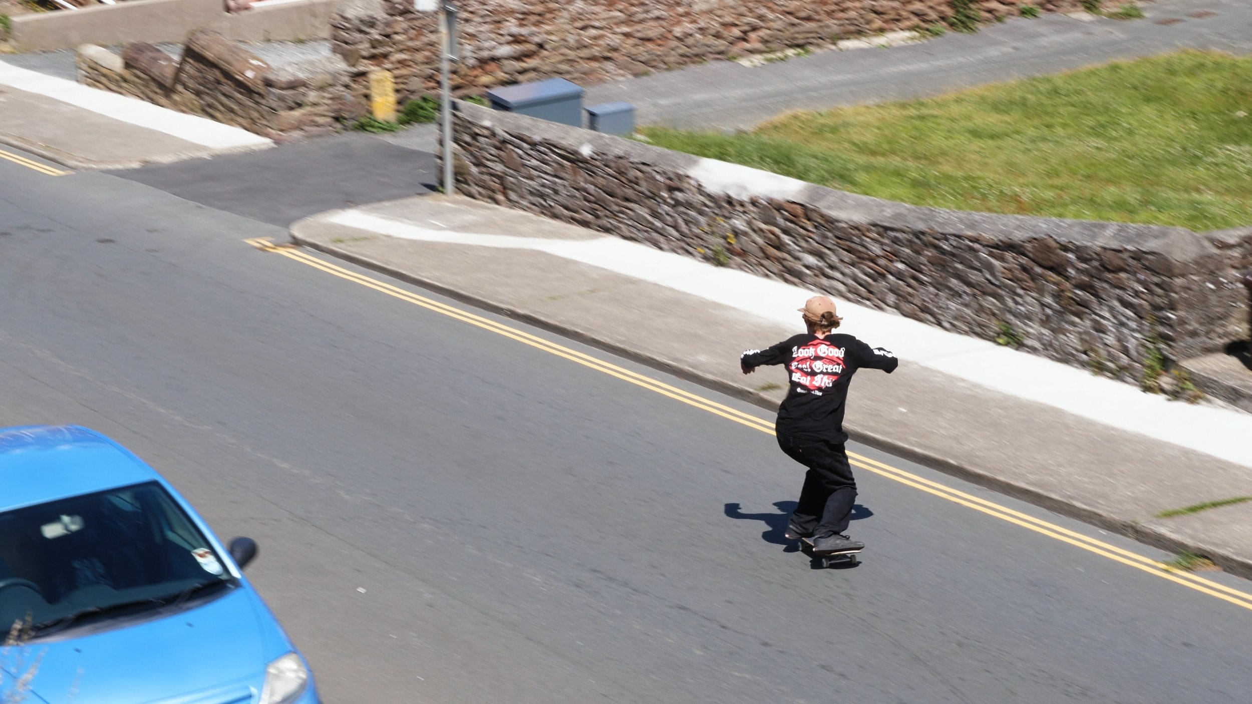 A person skating on a city street with a blue car passing by, residential area with stone walls and grass on the sidewalk.