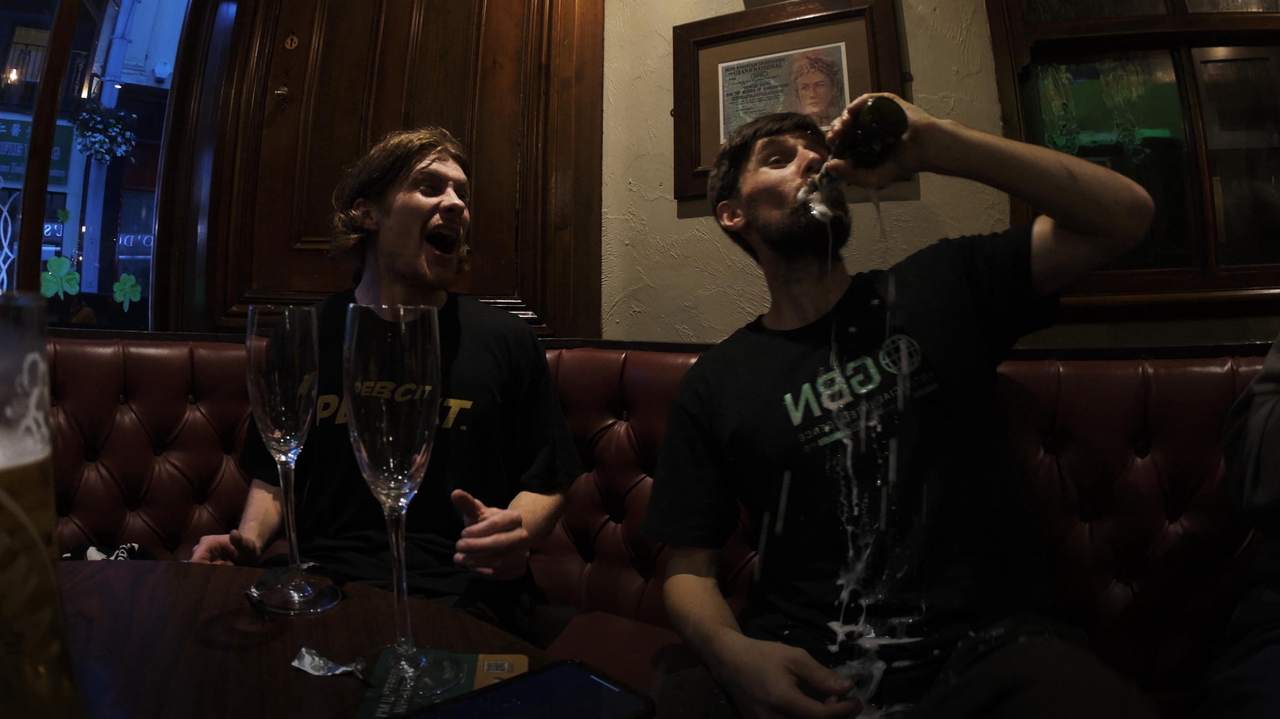 Two young men at a bar, one drinking beer and the other laughing, with empty champagne glasses on the table.
