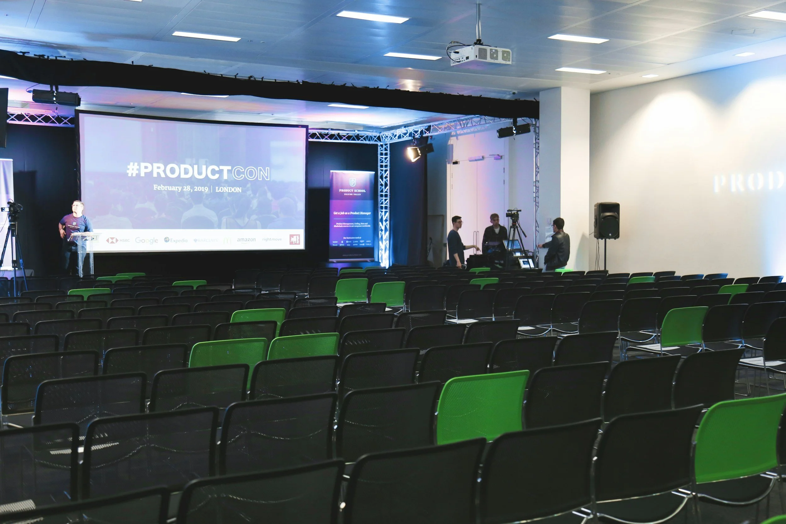 Empty conference room setup for ProductCon event, with rows of chairs facing a large screen displaying '#ProductCon' in London, dated February 28, 2019. People are setting up equipment near the stage.