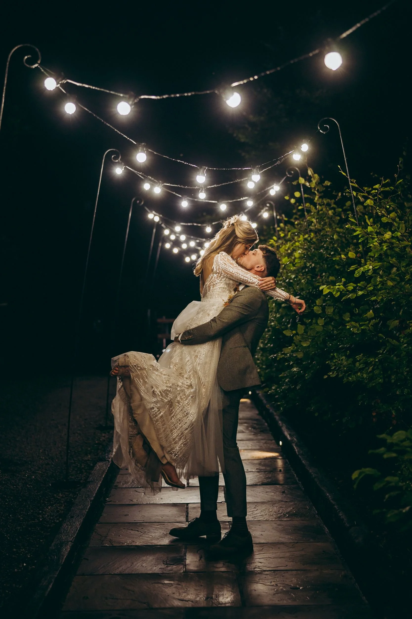 Romantic couple embracing under string lights at night, with man lifting woman in a wedding dress.