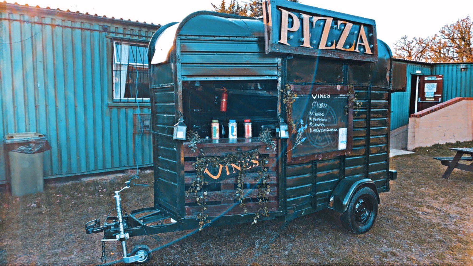 Food truck designed from a converted horse trailer with a "Pizza" sign, menu board, and condiment bottles, parked on grass near a teal metal building.