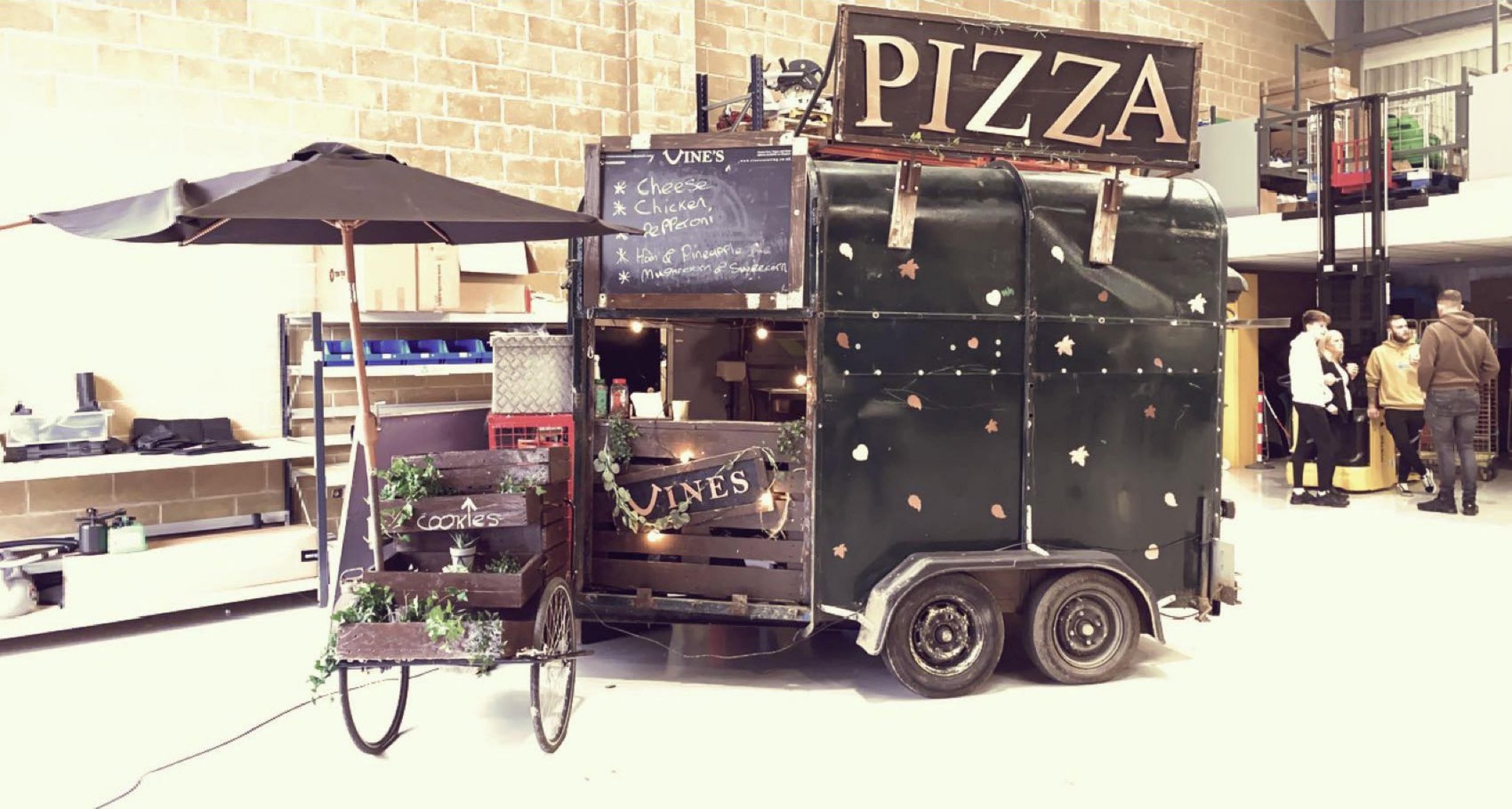Mobile pizza food truck indoors with a black umbrella shade, menu board displaying pizza options, and decorative plants, in a warehouse-like setting with a group of people in the background.