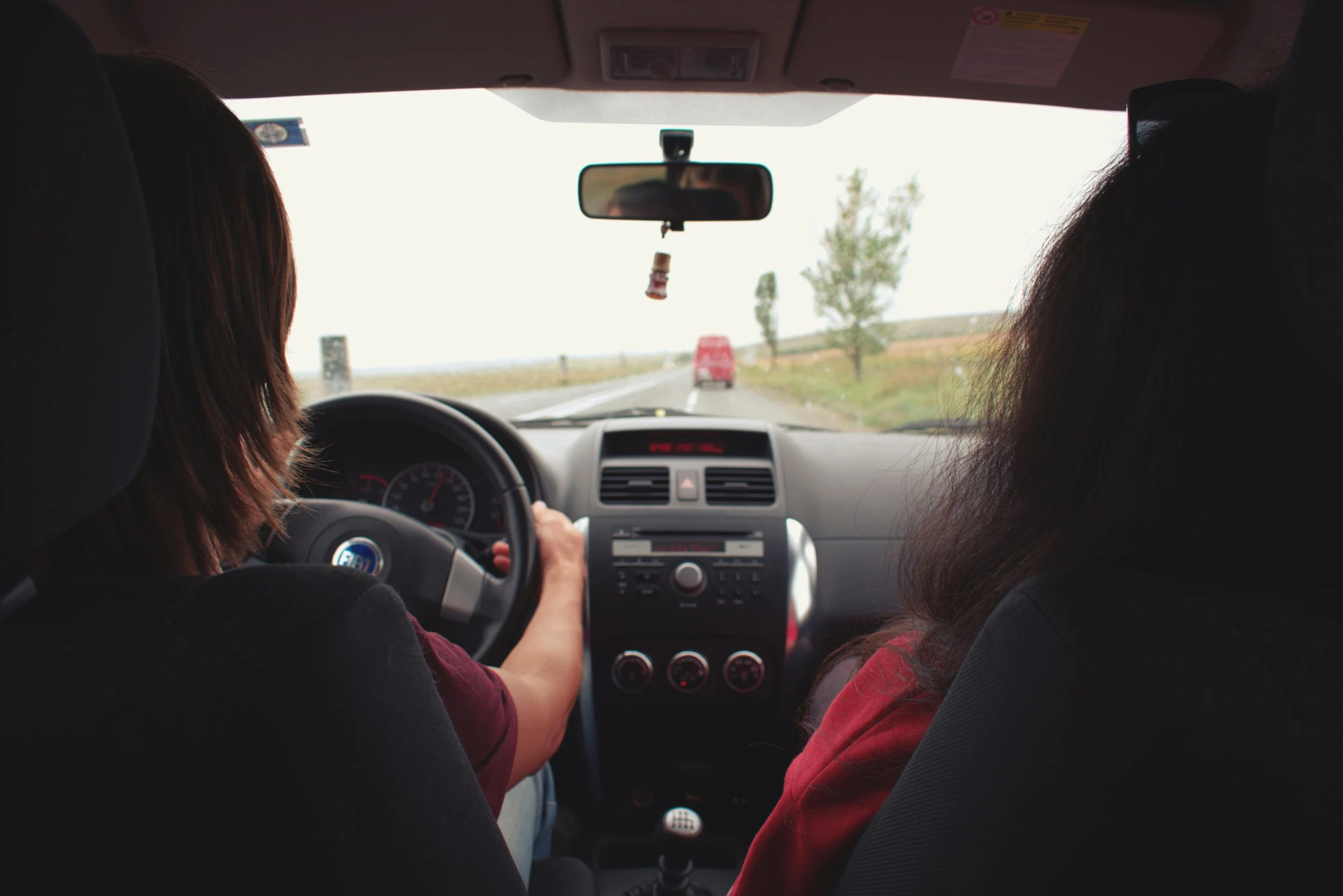 View from the back seat of a car showing a young woman driving while another person sits in the passenger seat, looking out at the road ahead through the windshield.