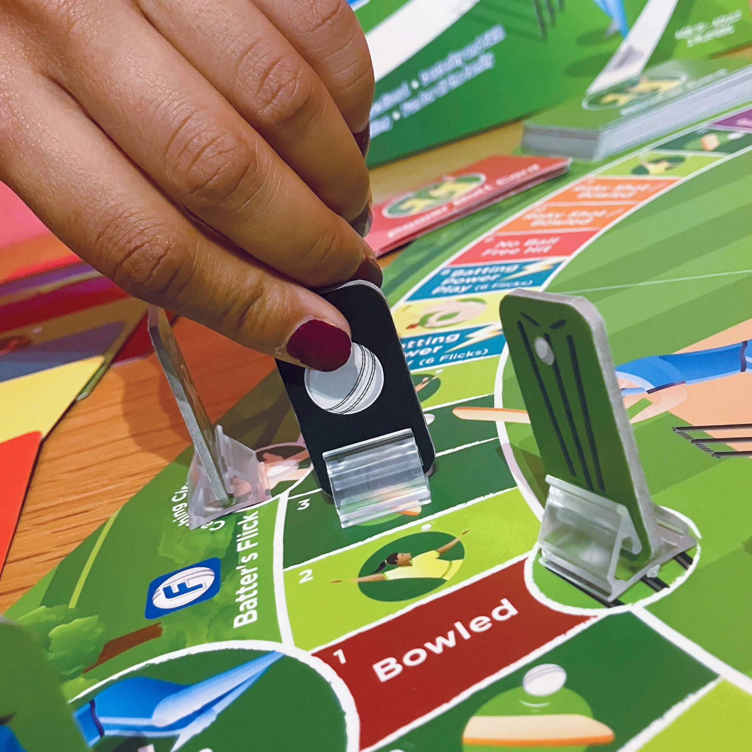 Close-up of a person's hand reaching for a game piece on a colourful game board, with Square labelled 'Bowled' visible.