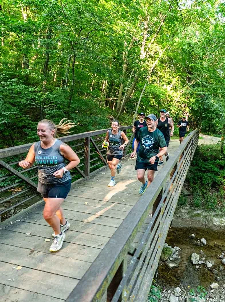 Group of seven runners jogging on a wooden bridge through a lush green forest during daytime.