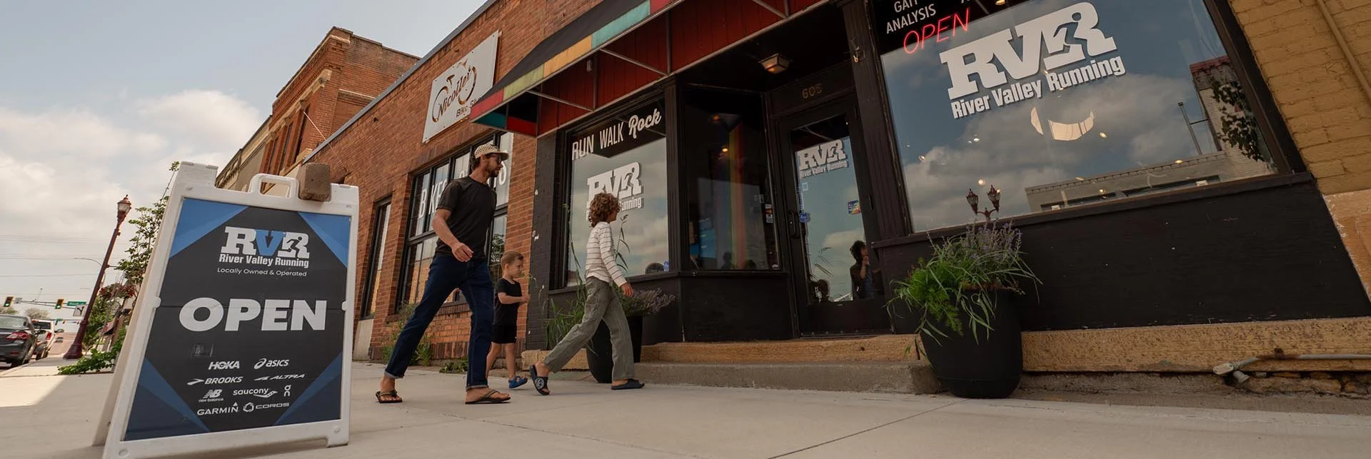 People walking past a storefront with a large window and signs for River Valley Running, including an open sign and logos for various athletic brands. There is a sidewalk sign with the same information outside the store, and a small group, including children and adults, is walking by.
