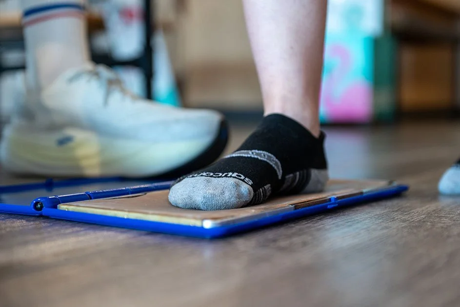 Close-up of a person standing on a foot measurement device, with a white athletic shoe and another shoe visible in the background.