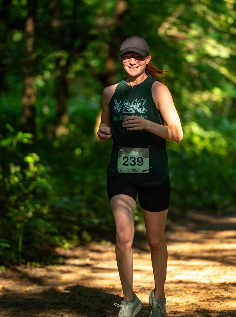 A woman jogging through a wooded trail while smiling, wearing black athletic clothes, a gray cap, sunglasses, and a race bib with the number 239.