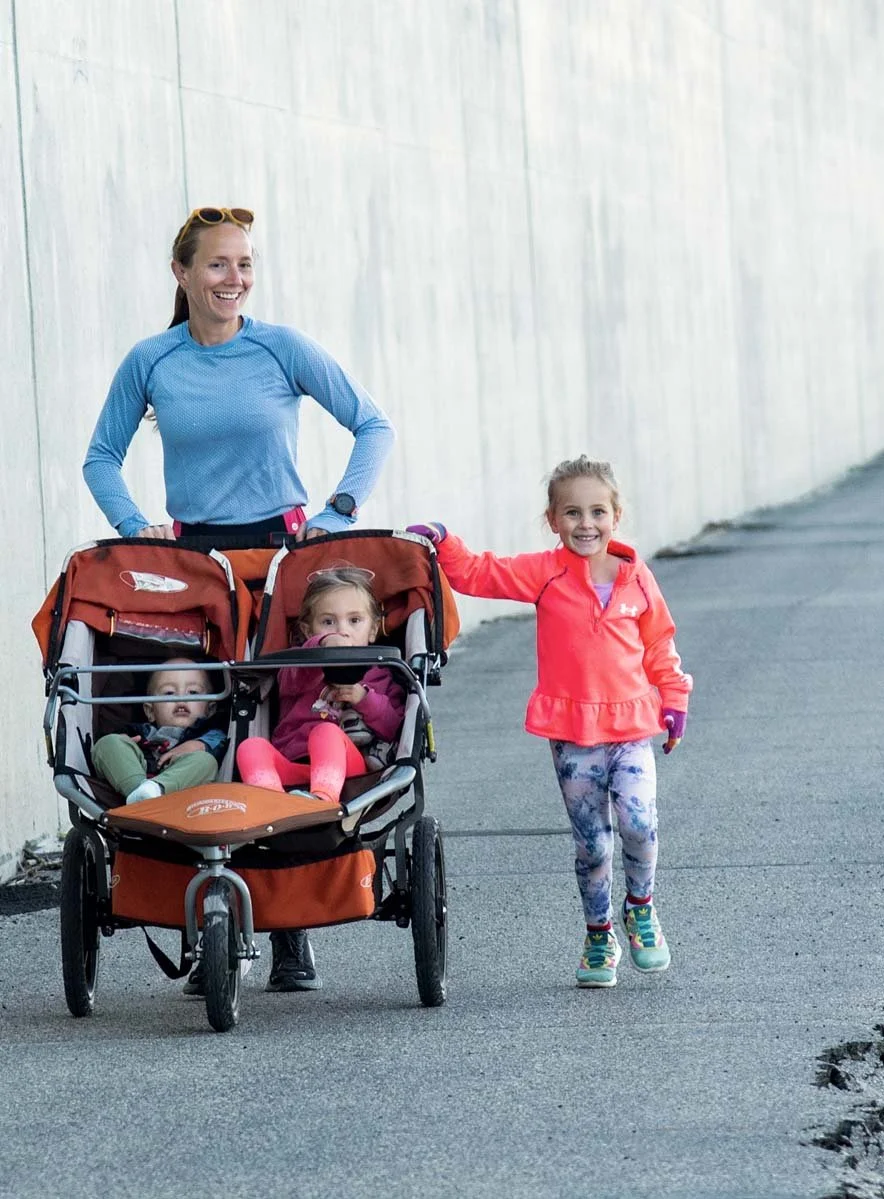 A smiling woman jogging with a baby stroller with two young children inside and another young girl walking alongside, touching the stroller, on a paved pathway next to a tall concrete wall.