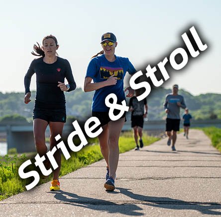 Two women running on a paved path outdoors, with several other people jogging in the background, on a clear day.