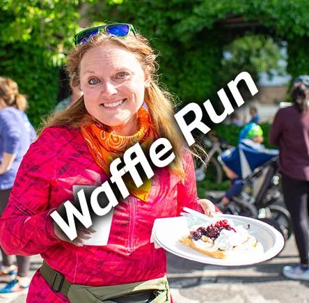 A smiling woman in a pink jacket holding a plate of waffles with berries, outdoors at a running event, with others in the background.