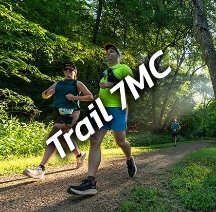 Two runners on a trail in a wooded area during a race, with a third runner in the distance.